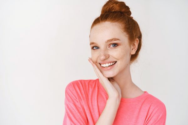 A red-headed woman with her hair in a bun smiles with her hand cupping the side of her jaw.