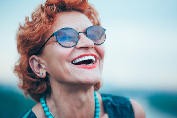 Older woman with red hair and sunglasses smiles through pink lipstick, showing off the results of her cosmetic dentistry procedures.